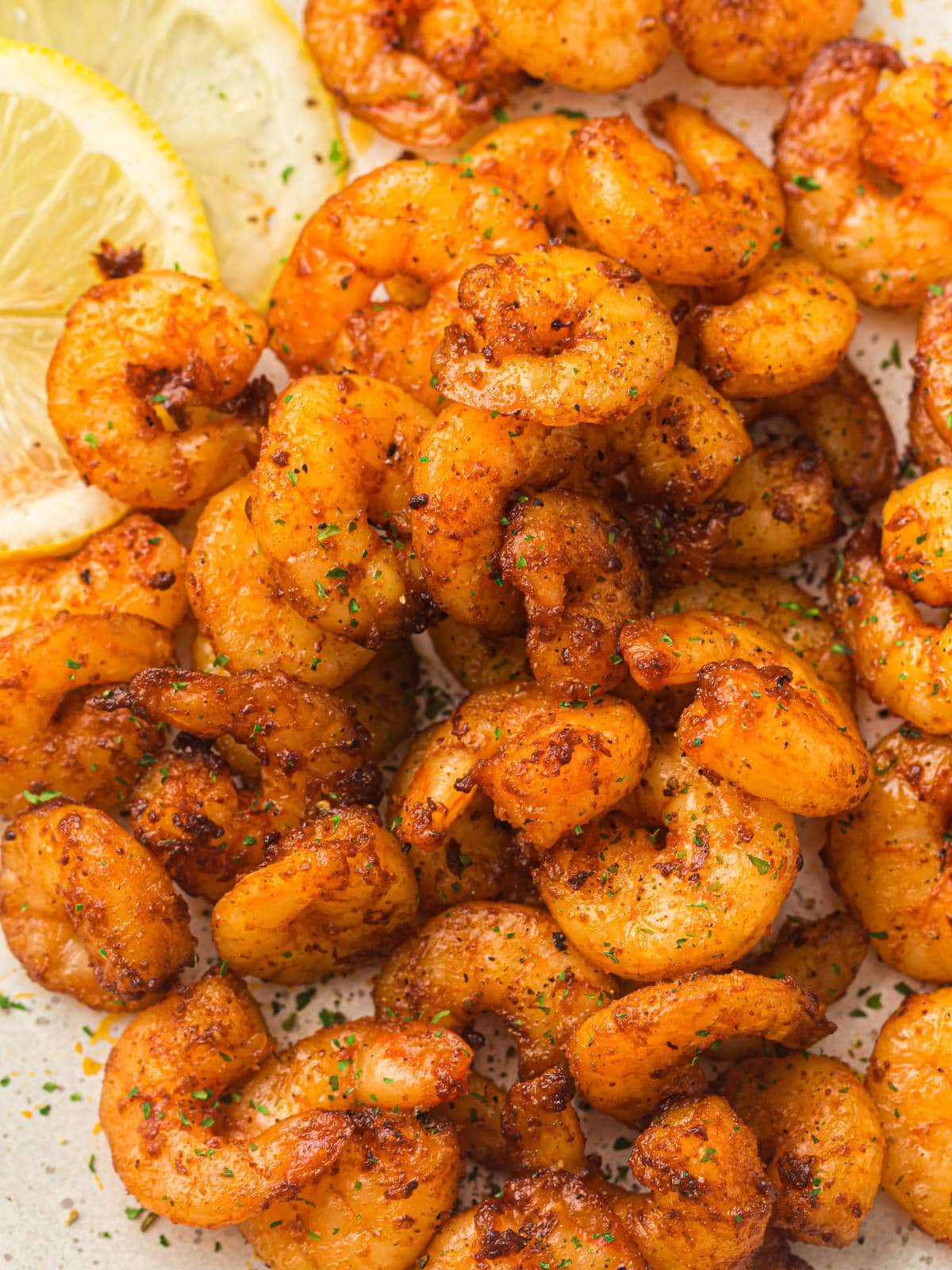 Seasoned cooked shrimp arranged on a plate with two slices of lemon in the corner, sprinkled with herbs.