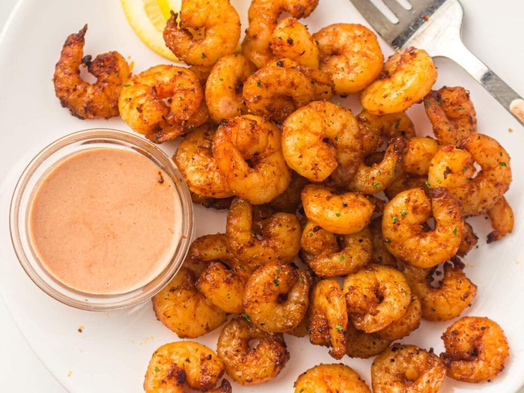 Seasoned cooked shrimp are arranged on a white plate with a small glass container of dipping sauce, a lemon wedge, and a fork.