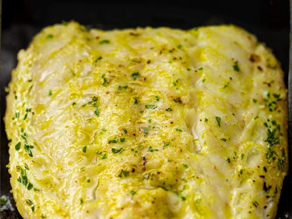 Close-up of a seasoned, baked white fish fillet with visible herbs and a golden, slightly crispy surface.