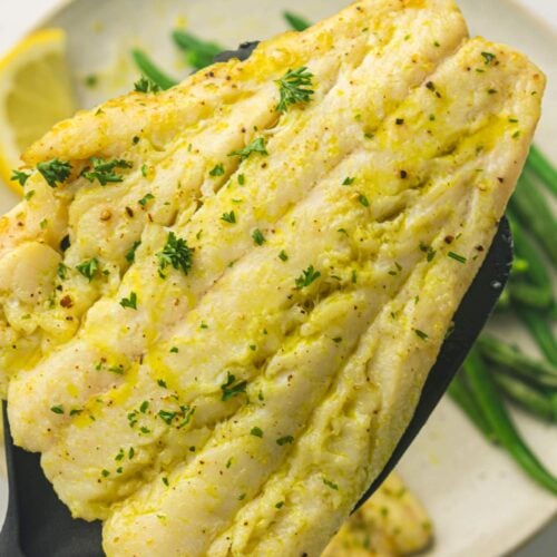 A cooked white fish fillet garnished with herbs is held on a black spatula above a plate with green beans and a lemon wedge.
