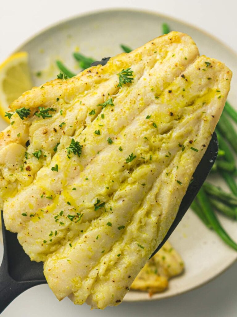 A cooked white fish fillet garnished with herbs is held on a black spatula above a plate with green beans and a lemon wedge.