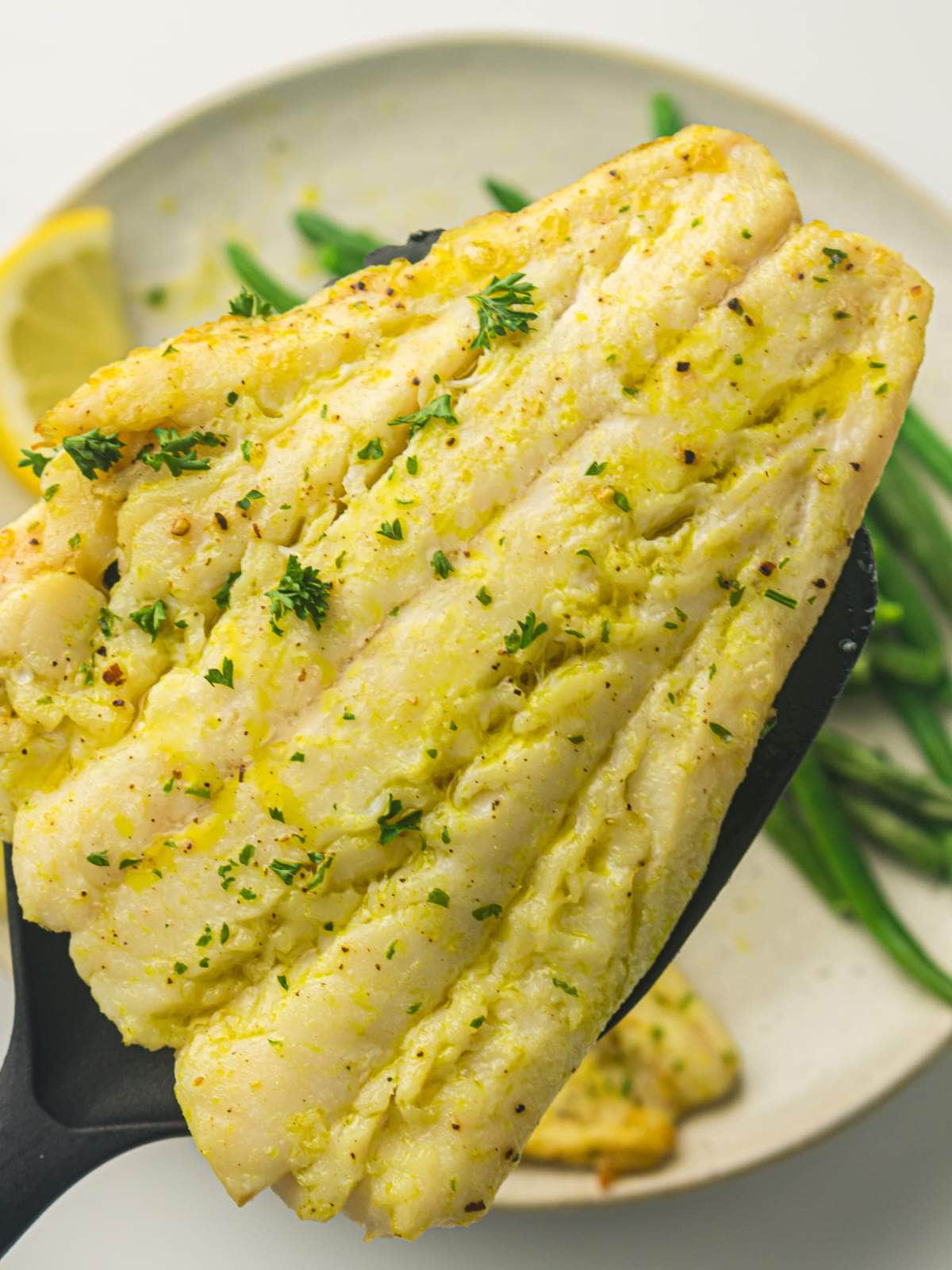 A cooked white fish fillet garnished with herbs is held on a black spatula above a plate with green beans and a lemon wedge.
