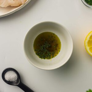 A white bowl with olive oil and chopped herbs, a lemon half, a black spoon with salt, and a plate with raw fish on a white surface.