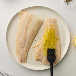 Two raw fish fillets on a plate, with one fillet being brushed with oil and seasoning using a black brush.