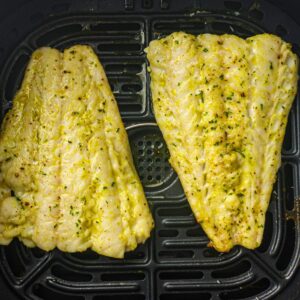 Two seasoned white fish fillets are cooking in an air fryer basket, with visible herbs and spices on the surface.