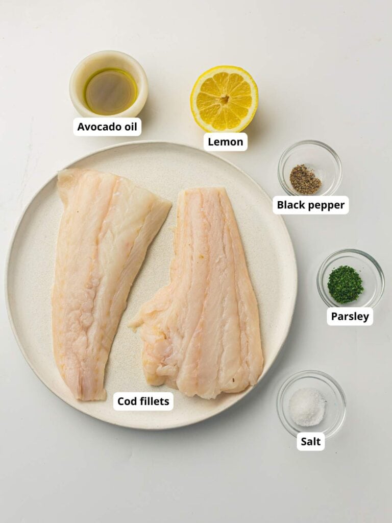 Plate with two raw cod fillets, surrounded by small bowls of avocado oil, lemon, black pepper, parsley, and salt, all labeled.