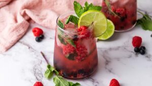 A glass of raspberry mocktail with mint leaves, lime slices, and ice on a marble surface, garnished with fresh berries and mint.