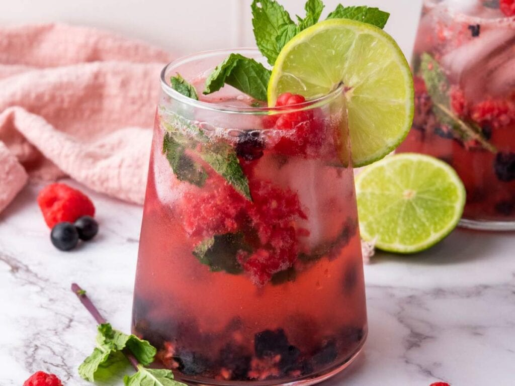 A glass of berry and mint cocktail with ice, garnished with a lime slice and fresh mint, sits on a marble surface with scattered berries and a sliced lime nearby.
