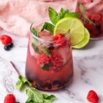 A glass of berry mocktail garnished with lime slices, mint leaves, raspberries, and blueberries on a marble surface.