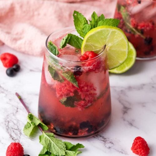 A glass of berry mocktail garnished with lime slices, mint leaves, raspberries, and blueberries on a marble surface.