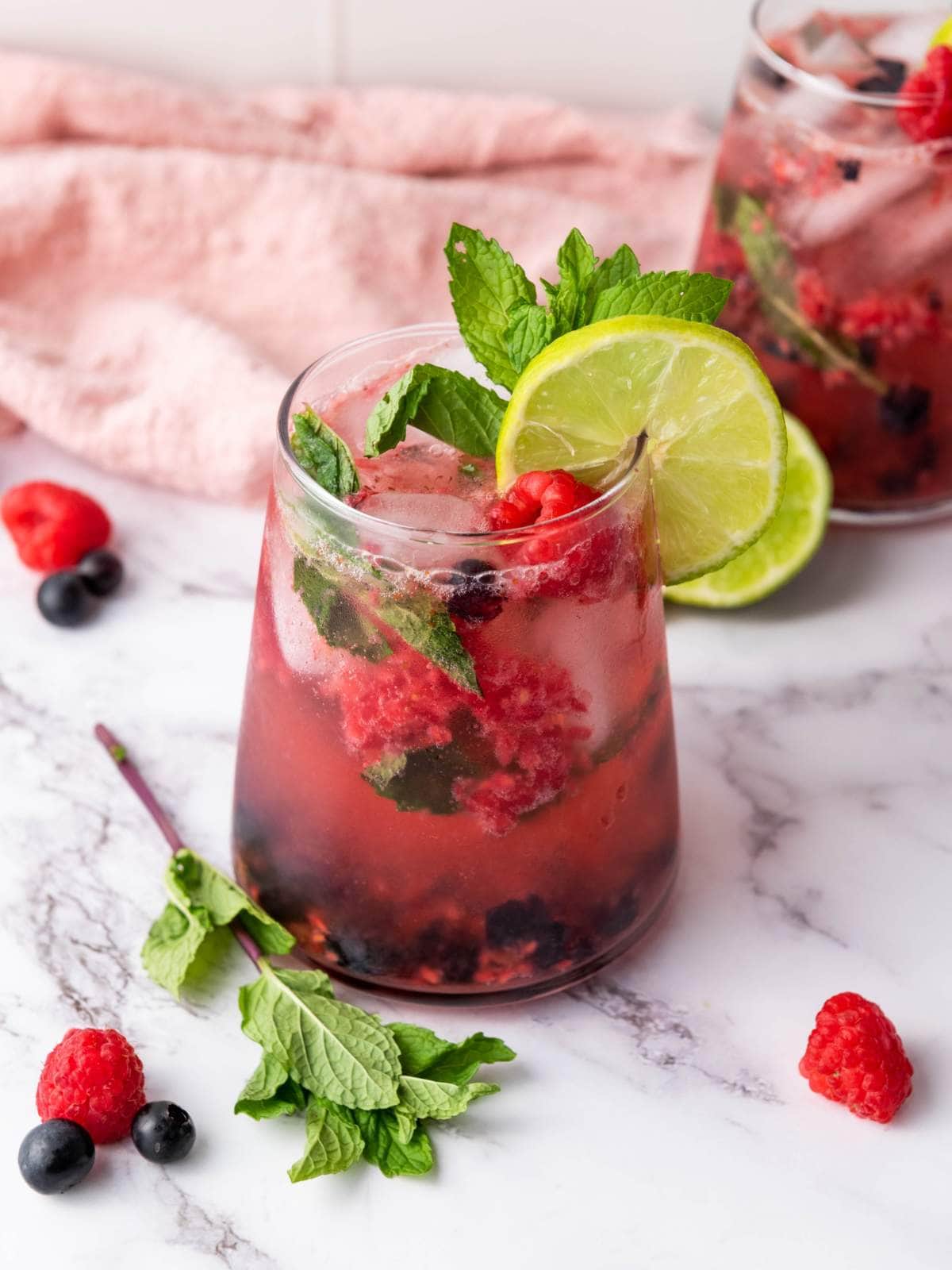 A glass of berry mocktail garnished with lime slices, mint leaves, raspberries, and blueberries on a marble surface.