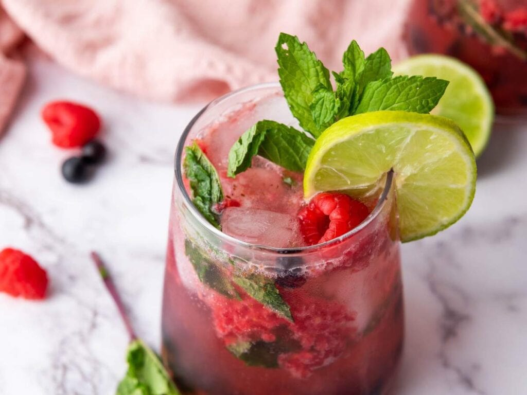 A glass of raspberry mocktail garnished with fresh mint leaves, raspberries, and two lime slices sits on a marble surface with scattered berries and mint nearby.