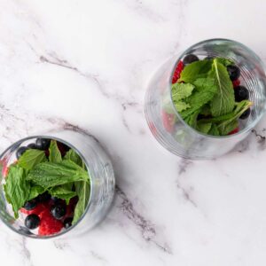 Two clear glasses with fresh raspberries, blueberries, and mint leaves on a white marble surface, viewed from above.