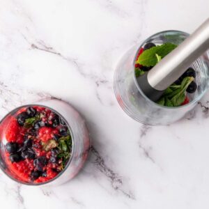 Two glasses on a marble surface, one with muddled berries and mint, the other with whole berries, mint, and a muddler inside.