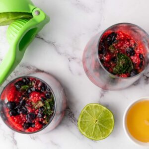 Two glasses with mixed berries and mint, a green citrus squeezer, half a lime, and a small bowl of juice on a marble surface.