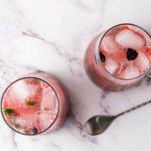 Two round glasses filled with pink iced drinks, blackberries, mint, and ice cubes sit on a marble surface next to a metal spoon.