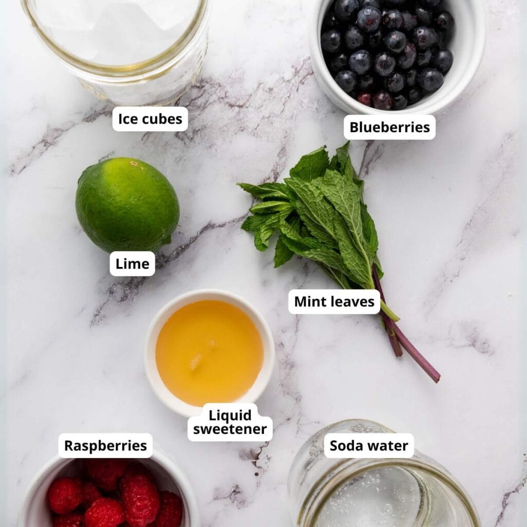 A collection of ingredients on a marble surface: ice cubes, blueberries, lime, mint leaves, raspberries, liquid sweetener, and soda water, each labeled.
