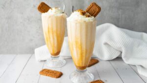 Two tall glasses of creamy Biscoff milkshake topped with whipped cream and biscuits, with extra biscuits on a white wooden surface and a white cloth in the background.
