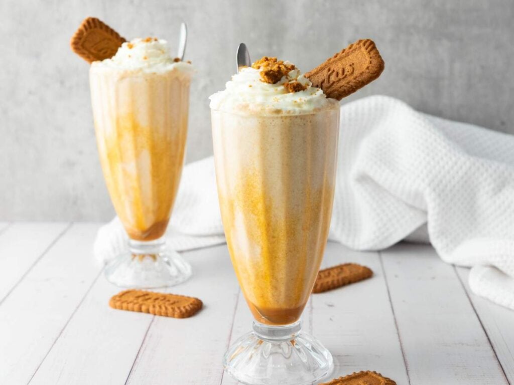 Two tall glasses of creamy milkshake topped with whipped cream and a Biscoff cookie, set on a white wooden surface with extra cookies and a white textured cloth in the background.