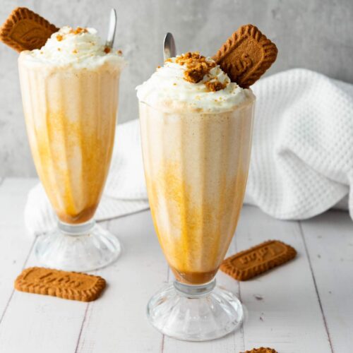 Two tall glasses of milkshake with whipped cream, crushed cookies, and whole biscuit garnishes, set on a white surface with scattered cookies and a textured white cloth in the background.