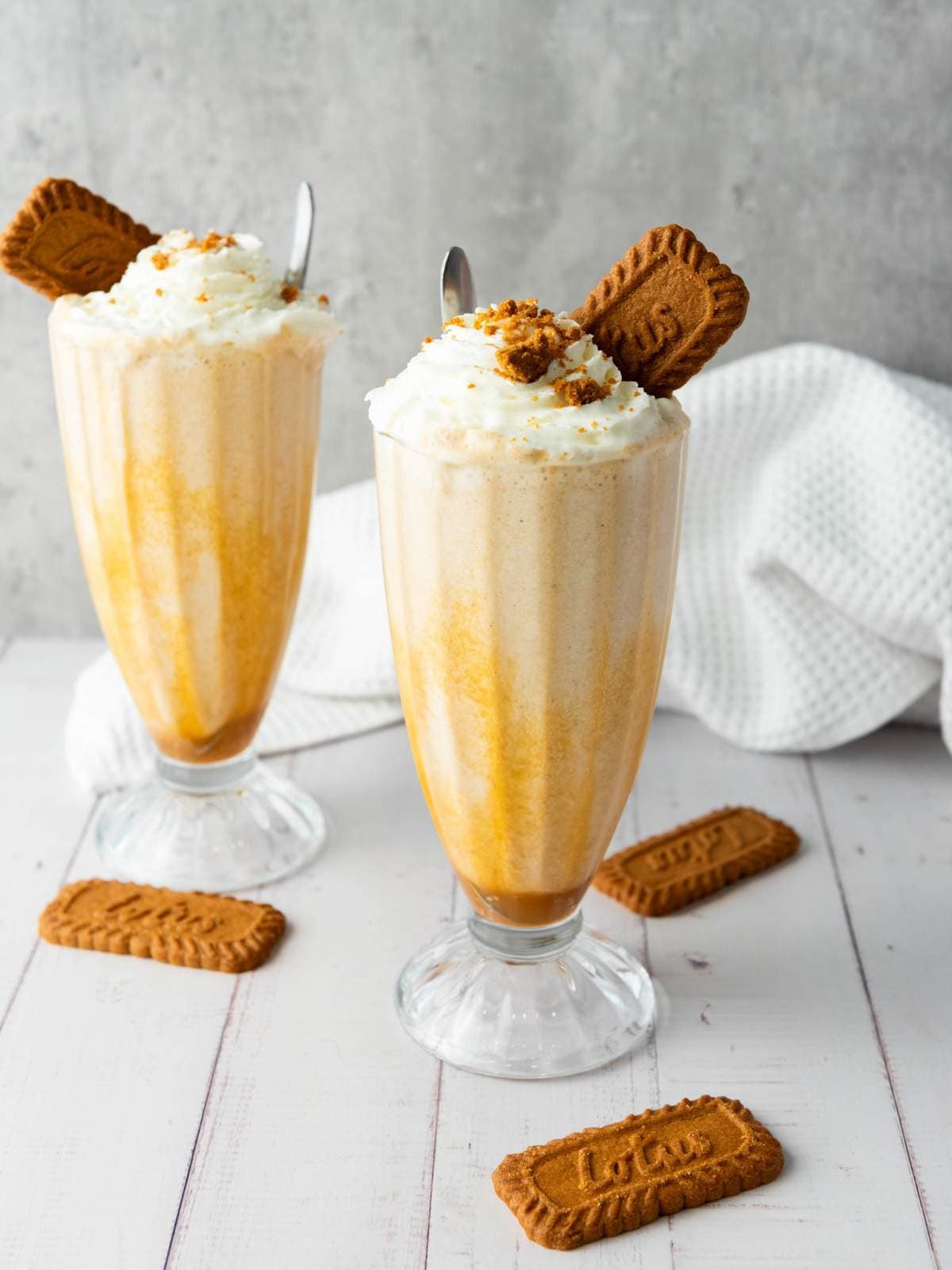 Two tall glasses of milkshake with whipped cream, crushed cookies, and whole biscuit garnishes, set on a white surface with scattered cookies and a textured white cloth in the background.