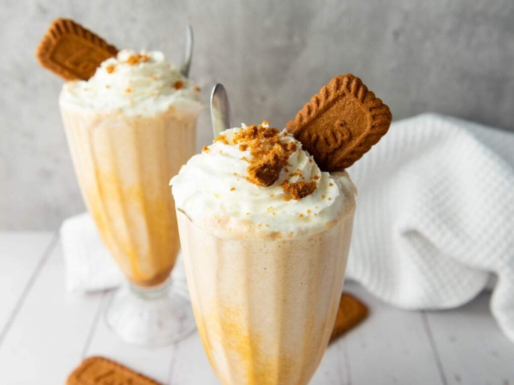 Two tall milkshakes topped with whipped cream and cookie crumbles, each garnished with a whole cookie in a clear glass, on a light surface with a white towel in the background.