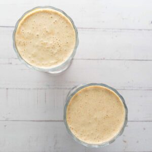 Two glass cups filled with a thick, light brown smoothie or milkshake are placed on a white wooden surface, viewed from above.