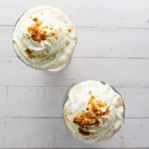 Two milkshakes topped with whipped cream and crumbled cookie pieces, viewed from above on a white wooden surface.