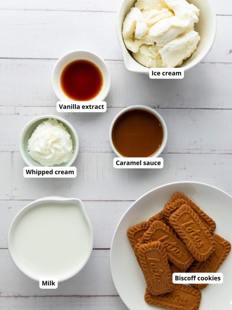 Five labeled bowls contain ice cream, vanilla extract, whipped cream, caramel sauce, milk, and Biscoff cookies arranged on a white wooden surface.