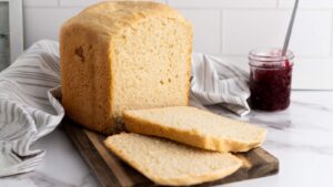 A loaf of bread with two slices cut sits on a wooden board next to a jar of red jam and a striped cloth on a marble counter.