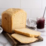 A loaf of bread with two slices cut sits on a wooden board; a jar of red jam with a spoon is in the background on a marble countertop.