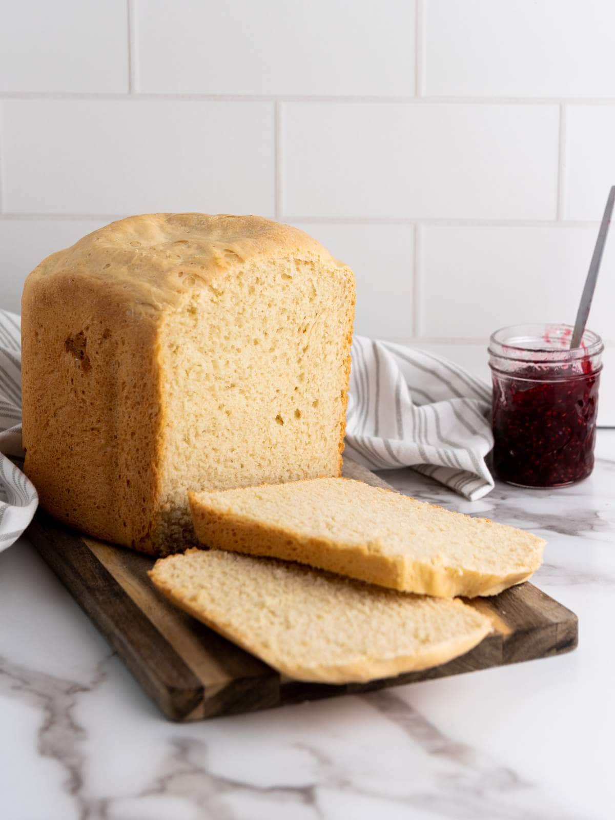 A loaf of bread with two slices cut sits on a wooden board; a jar of red jam with a spoon is in the background on a marble countertop.