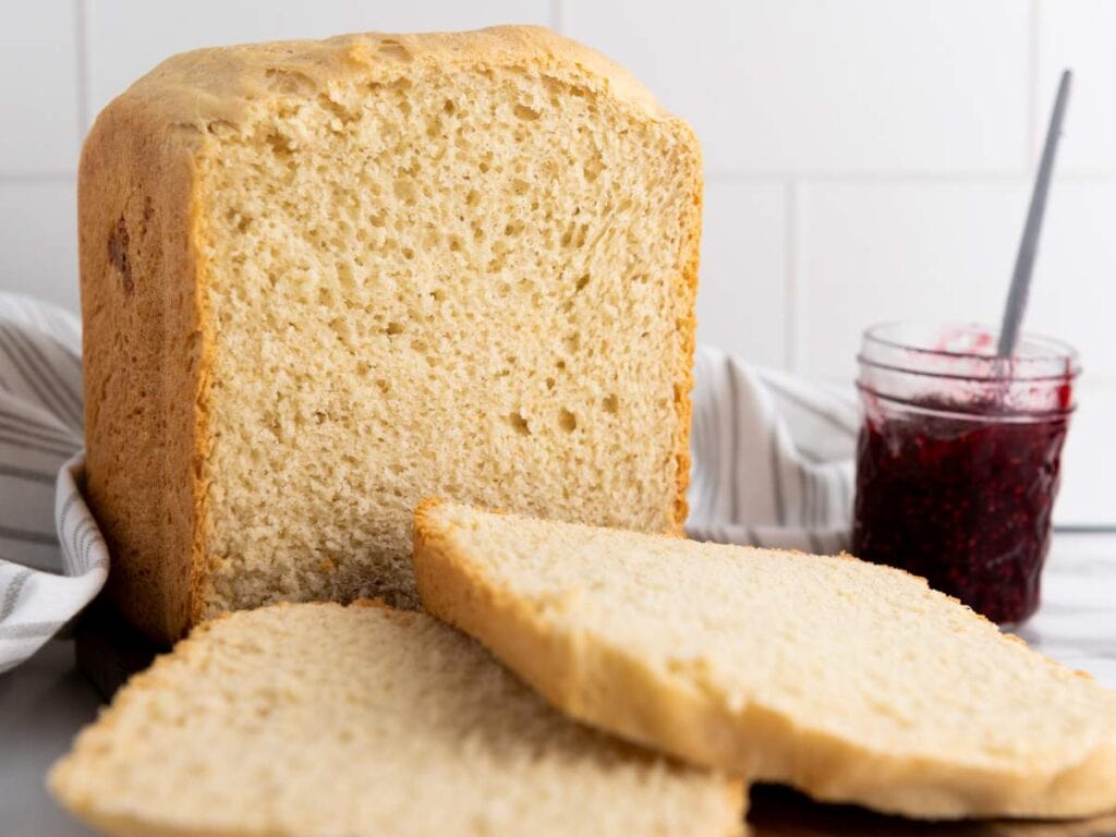 A loaf of homemade bread with two slices cut, placed on a surface next to a jar of red jam and a spoon.