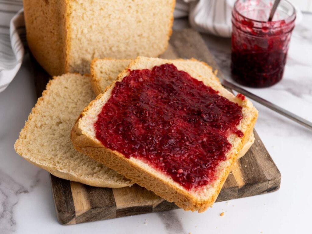 A slice of white bread with raspberry jam spread on top sits on a wooden board, with a loaf of bread and a jar of jam in the background.