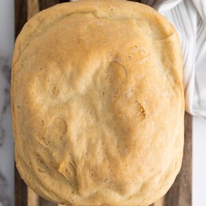A loaf of freshly baked bread with a golden-brown crust sits on a wooden cutting board, partially visible on the sides.