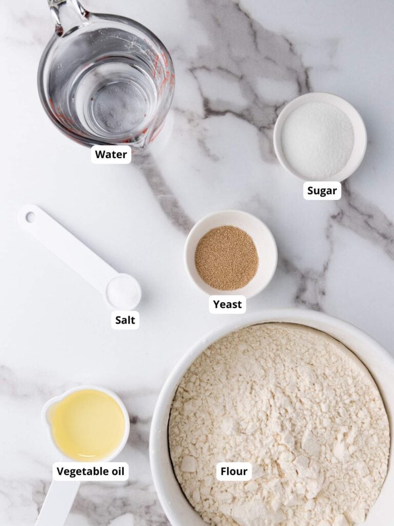 Measuring cups and bowls with labeled ingredients: water, sugar, yeast, salt, vegetable oil, and flour on a marble surface.