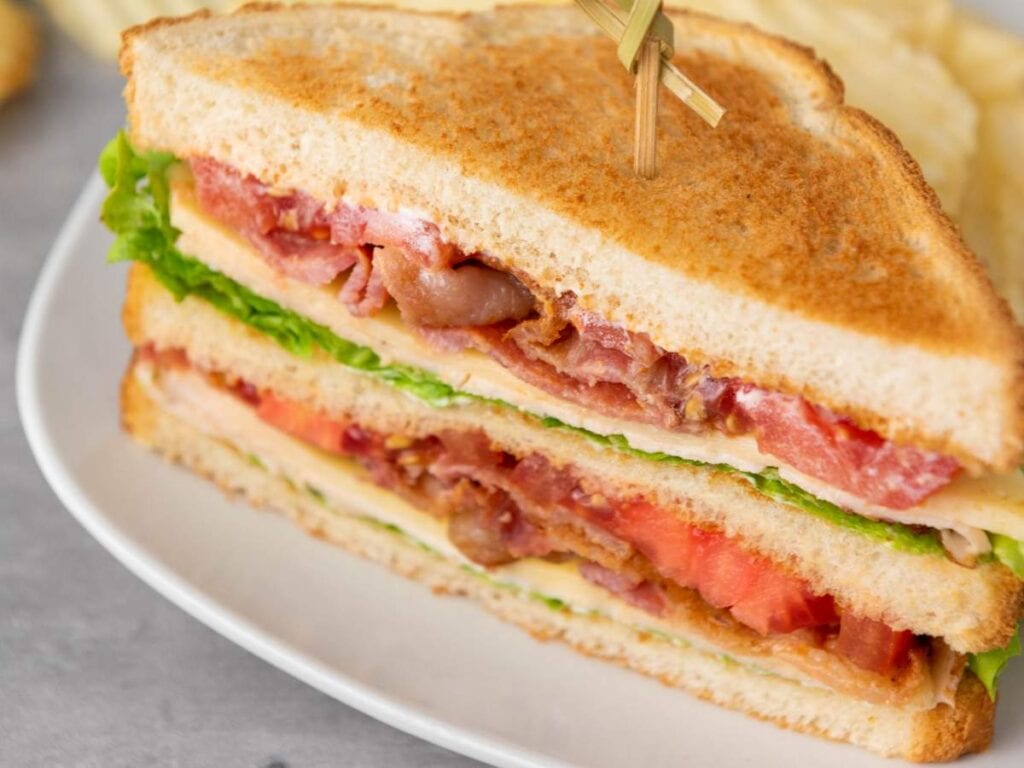 A BLT sandwich cut in half, stacked with bacon, lettuce, and tomato between toasted bread slices, served on a white plate.