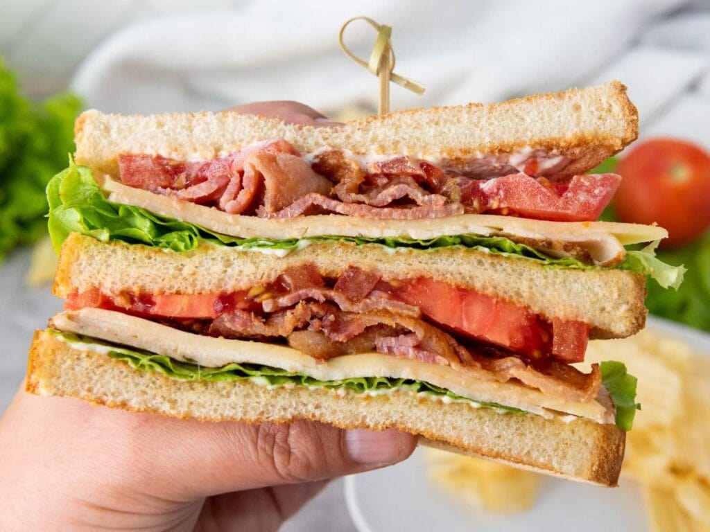 A hand holds a BLT sandwich with layers of bacon, lettuce, tomato, and mayonnaise between slices of toasted bread.