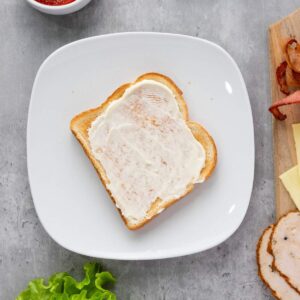 A slice of toasted bread with mayonnaise spread on a white plate, surrounded by sandwich ingredients on a gray surface.