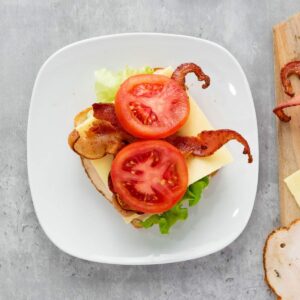 An open-faced sandwich with lettuce, cheese, bacon, and tomato slices on a white plate, viewed from above.
