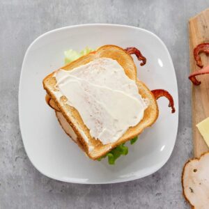 A sandwich on toasted bread with lettuce, bacon, turkey slices, and mayonnaise sits on a white plate; extra bacon and cheese are on a wooden board beside it.