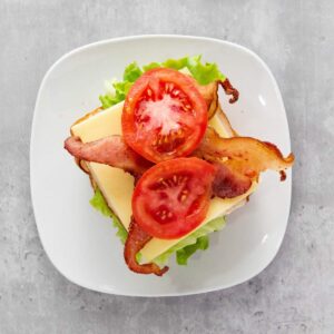 Open-faced sandwich with lettuce, cheese, two slices of bacon, and two tomato slices on white bread, served on a white plate against a gray background.