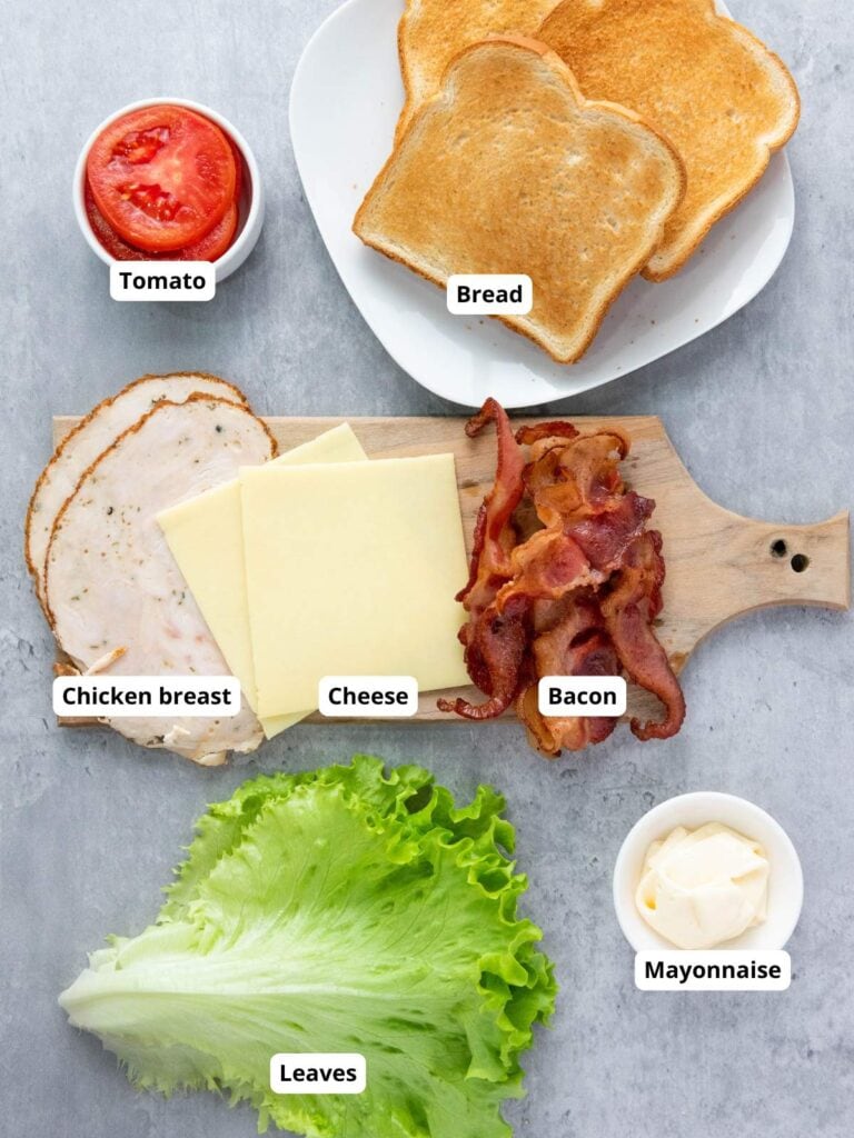 Sandwich ingredients arranged on a table: sliced tomato, toasted bread, chicken breast, cheese, bacon, lettuce leaves, and a small dish of mayonnaise. Each item is labeled.