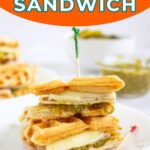 A turkey pesto croffle sandwich is stacked on a white plate, secured with toothpicks; more sandwiches and a jar of pesto are in the background.