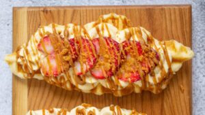 A waffle topped with whipped cream, sliced strawberries, crumbled cookie, and caramel drizzle on a wooden board.