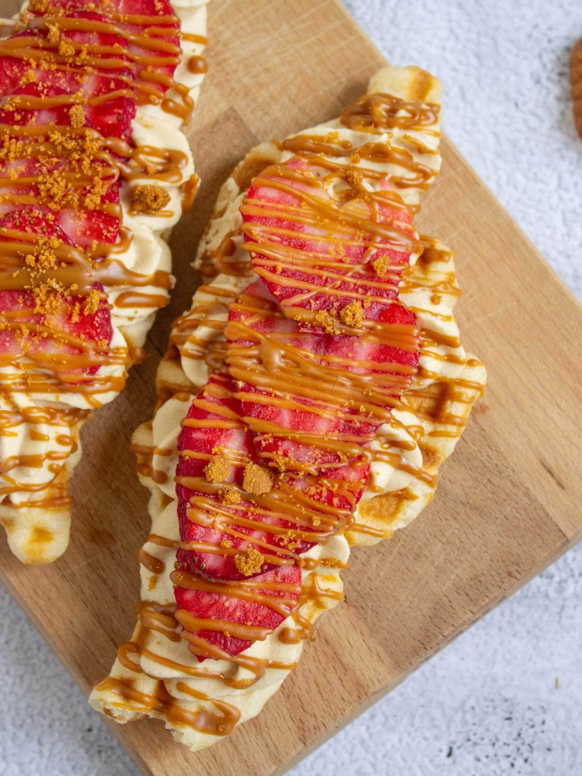 Two waffles topped with sliced strawberries, cream, crumbled cookie pieces, and a drizzle of caramel sauce on a wooden board.