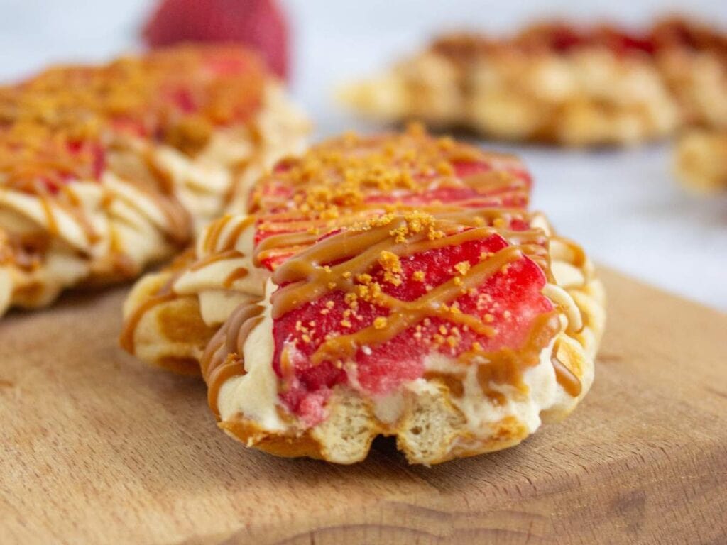 A close-up of a pastry topped with pink icing, caramel drizzle, and crumbs, placed on a wooden surface with more pastries in the background.