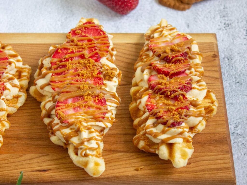 Two waffle sticks topped with cream, sliced strawberries, crumbled cookie, and caramel drizzle on a wooden board.