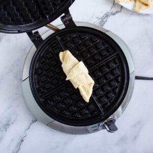 Uncooked crescent roll dough placed on the center of an open waffle maker, ready to be cooked.