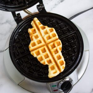 A partially cooked waffle in an irregular shape sits on the grid of an open waffle maker on a marble countertop.
